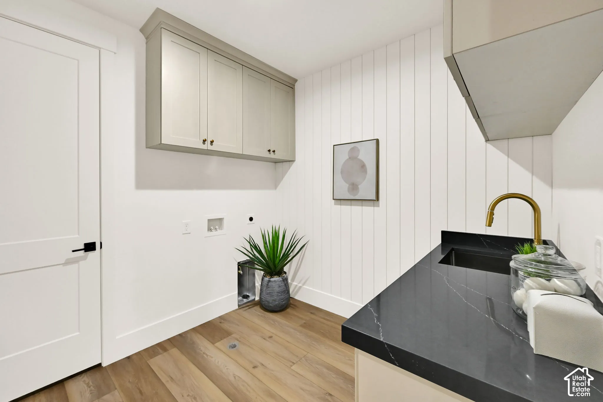 Wood-finish flooring, white shiplap accent wall, dark countertop with integrated sink, brass-finish faucet, and light-colored cabinetry
