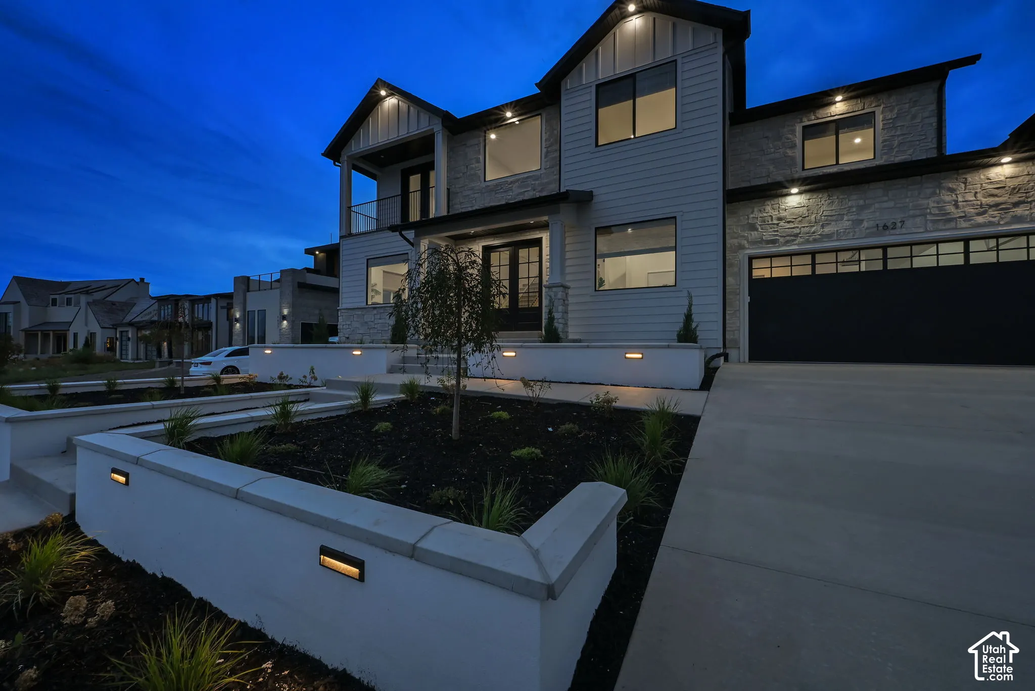 Modern exterior with stone and siding facade, integrated landscape lighting, a concrete driveway, a two-car garage, and a second-story balcony with a glass railing