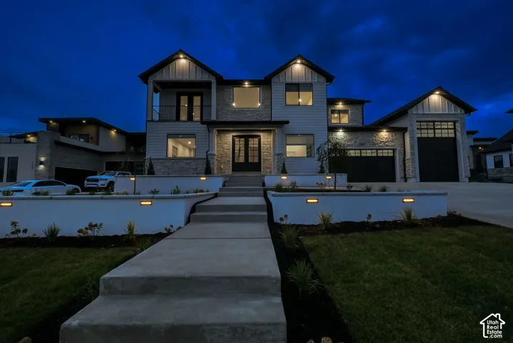 Contemporary exterior featuring board and batten siding, stone accents, multiple garages, a prominent balcony, and an illuminated concrete pathway