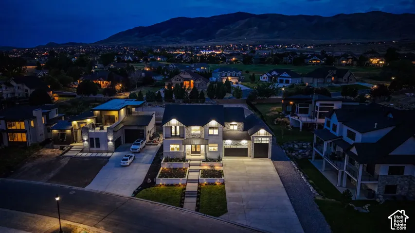 Modern architectural residence showcasing illuminated interiors with geometric facade elements, expansive paved driveway, manicured landscaping with integrated hardscaping, and mountain backdrop visible from the property