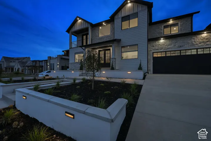 Modern exterior with stone and siding facade, integrated landscape lighting, a concrete driveway, a two-car garage, and a second-story balcony with a glass railing