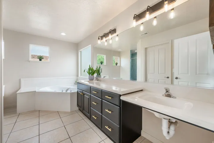 Built-in corner tub, dual vanities with dark cabinetry, integrated sinks, large wall-mounted mirror, and tile flooring
