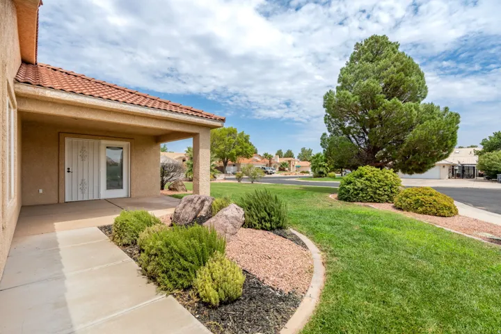 Covered front porch with tile flooring and white entry door opening to a manicured landscape featuring curved planting beds with desert shrubs, river rock accents, and mature trees