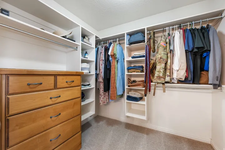 Spacious walk-in closet featuring custom shelving with built-in cubbies for organized storage, a wooden dresser with multiple drawers, dual clothing rods with hanging space for an extensive wardrobe, and neutral carpeting throughout