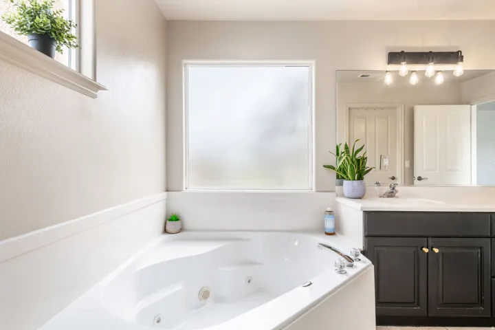 Built-in jetted tub with polished chrome fixtures, white countertop vanity with dark cabinetry, and an elongated frosted window