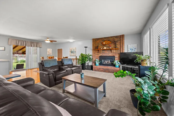Spacious living room featuring multiple seating areas with dark leather furnishings, wood-finish coffee table, brick fireplace with decorative surround, recessed ceiling lighting, large windows with plantation shutters, neutral carpet flooring, and abunda
