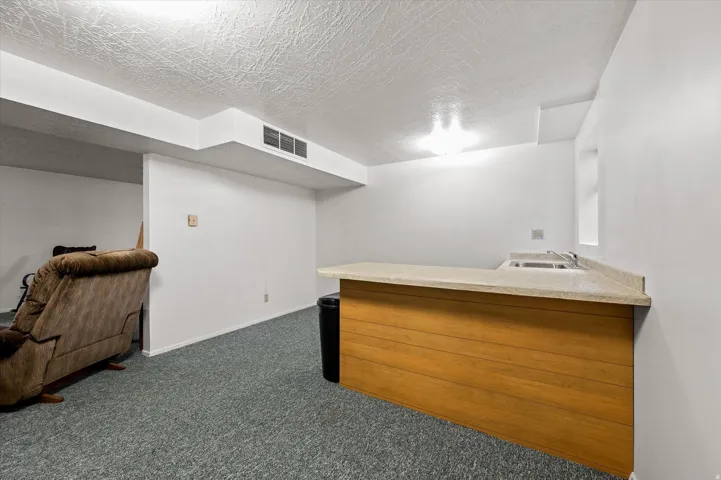 Finished basement featuring carpeted flooring, wood-finish cabinetry with countertop workspace, recessed ceiling lighting, and white drywall walls throughout the space