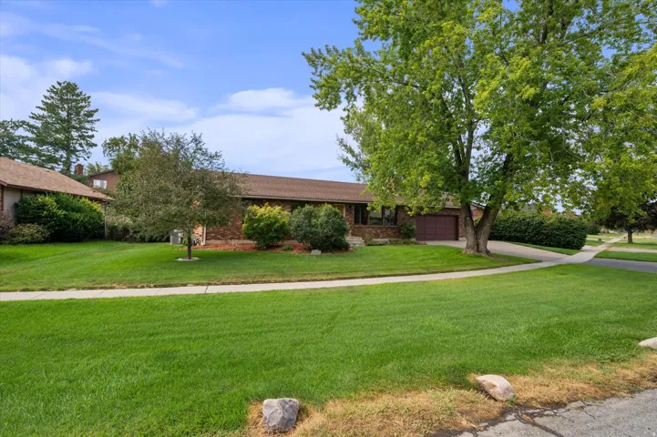 Brick exterior with a brown garage door, expansive green lawn, mature trees, and a concrete driveway