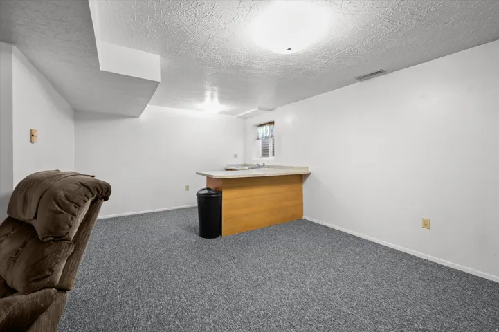 Finished basement recreation room featuring carpeted flooring, wood-finish counter with storage, recessed ceiling lighting, and a high small window for natural light