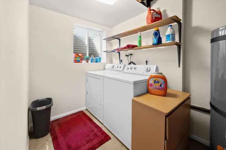 Efficient laundry room featuring white front-load washer and dryer, wood-finish shelving with ample storage for cleaning supplies, stainless steel trash receptacle, and wood-finish work surface for folding and sorting