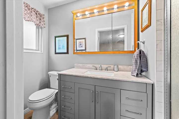 Spa-inspired bathroom featuring a gray vanity with multiple drawers and cabinet storage, white quartz countertop, and a large illuminated mirror with Hollywood-style lighting