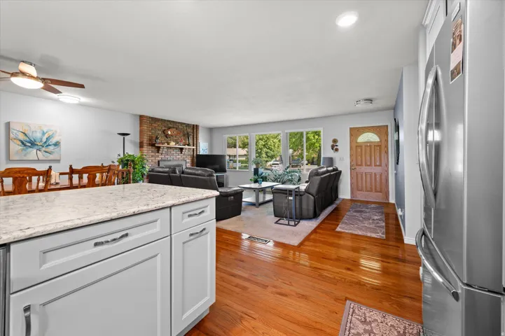 Open-concept kitchen and living area featuring white cabinetry with hardware accents, quartz countertops, stainless steel appliances including a French-door refrigerator, warm wood-finish flooring throughout, and abundant natural light from multiple glass