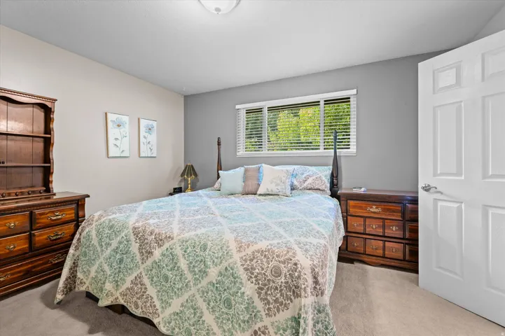 Serene primary bedroom showcasing wood-finish nightstands with multiple drawers, a four-poster bed frame, plantation shutters with natural light filtration, neutral carpeting, and soft gray wall tones complemented by crisp white trim and closet doors