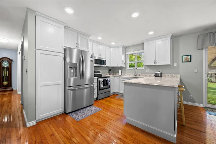 Modern kitchen featuring white shaker cabinetry, stainless steel appliances including a French-door refrigerator and dual ovens, granite countertops, a centered island with breakfast bar seating, recessed lighting, and warm wood-finish flooring throughout