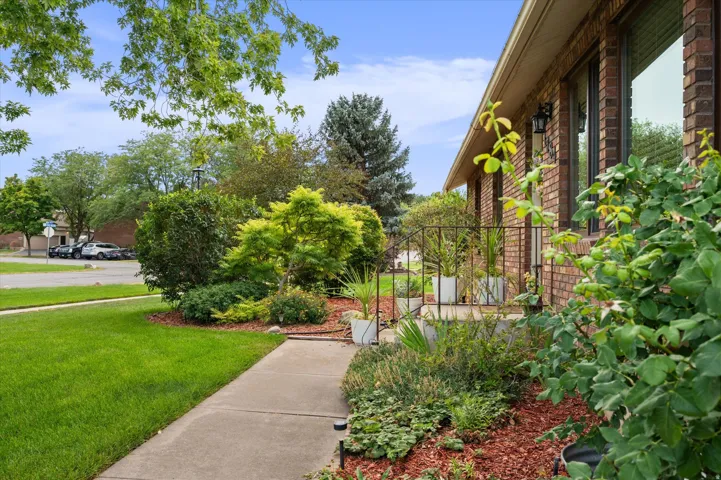 Brick exterior with multiple windows, concrete walkway, mature trees, and landscaped garden beds