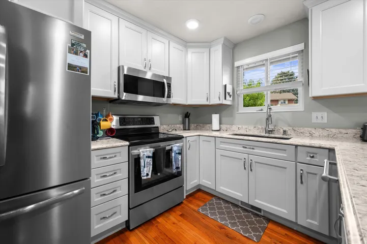 Contemporary kitchen featuring white shaker-style cabinetry, stainless steel appliances including a built-in microwave and electric range, granite countertops with undermount sink and chrome faucet, hardwood flooring, and abundant natural light from corne