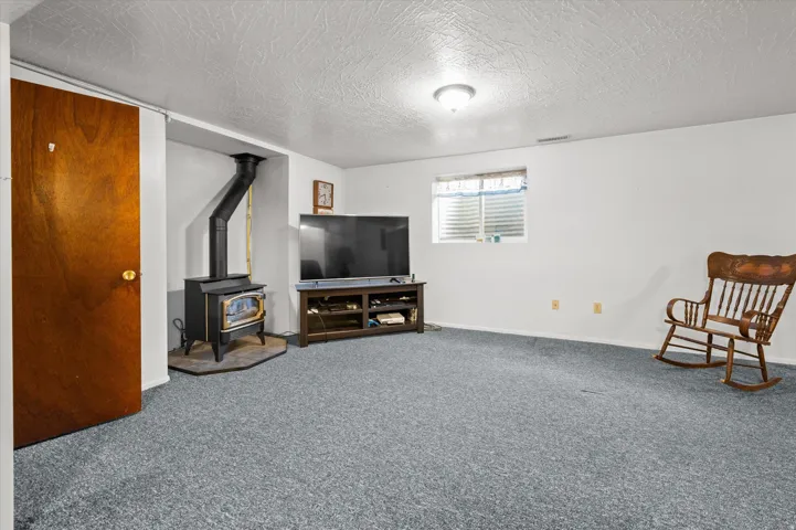 Spacious recreation room featuring wall-to-wall carpeting, a wood-burning stove with metal chimney, built-in media console, large picture window with blinds, and solid wood entry door