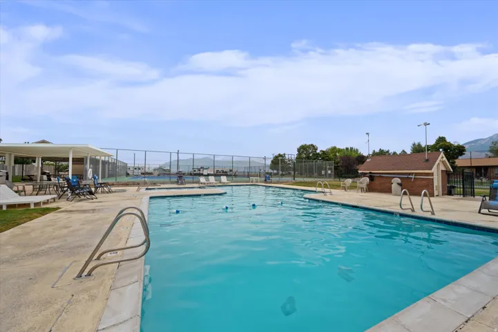 Outdoor swimming pool with concrete decking, surrounding chain-link fencing, a covered patio area, and a brick-sided building