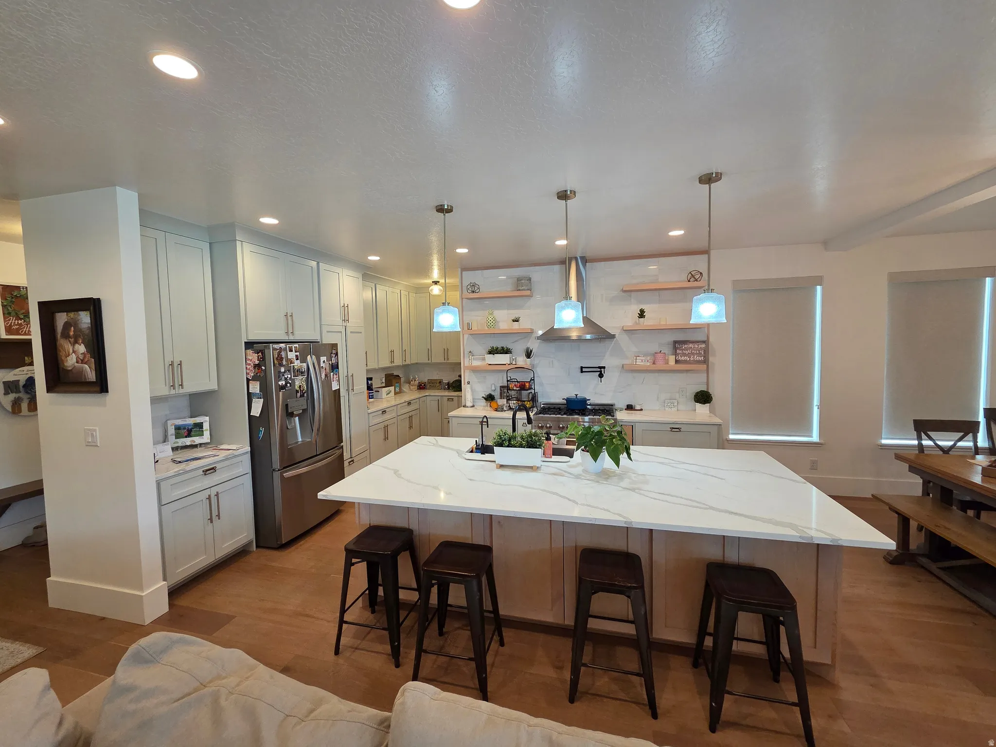 Modern kitchen featuring a large waterfall-edge island with quartz countertop, stainless steel French-door refrigerator, white cabinetry with hardware accents, pendant lighting, and wood-finish flooring throughout the open living space