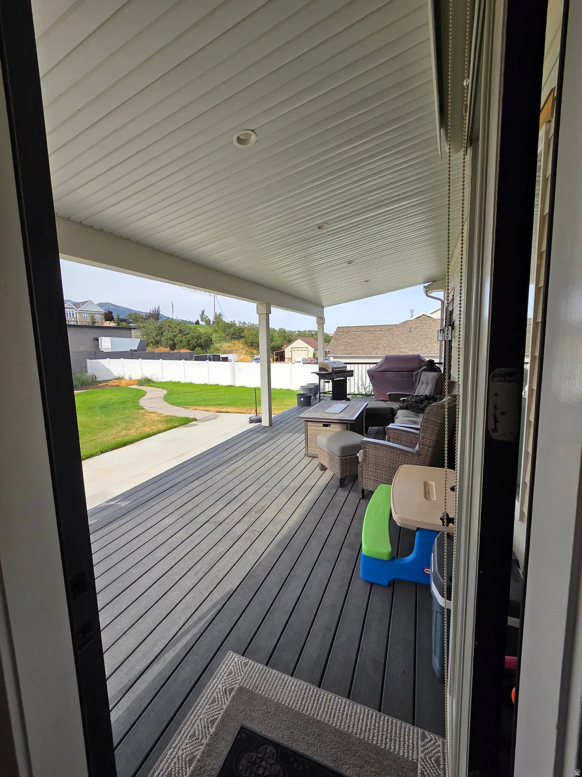 Expansive covered deck with recessed lighting and composite decking, extending to a manicured lawn with a concrete pathway and white fencing