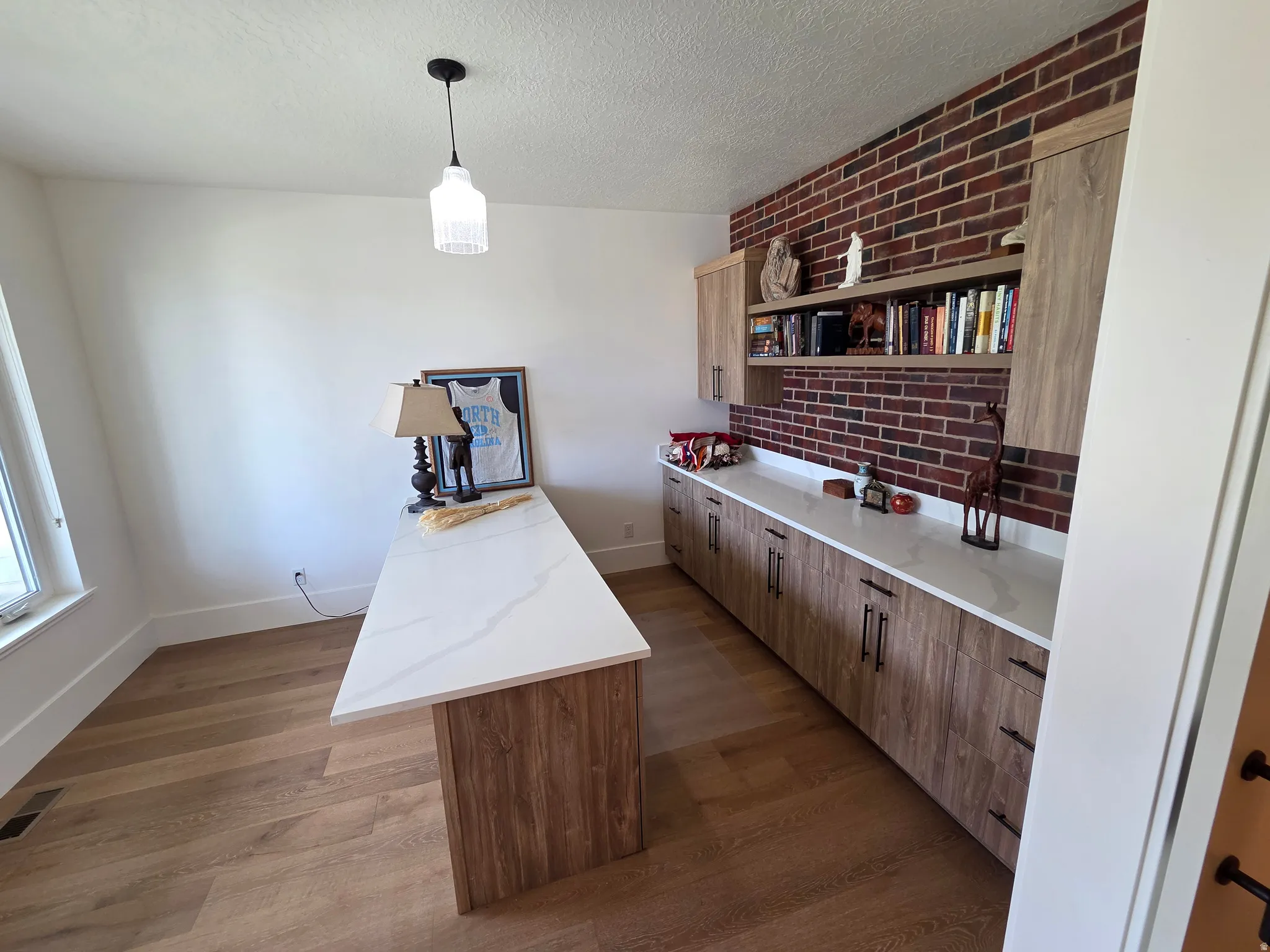 Modern home office featuring a wood-finish island countertop with ample workspace, exposed red brick accent wall with built-in shelving and cabinetry, polished concrete countertops, warm wood-finish cabinetry throughout, and pendant lighting