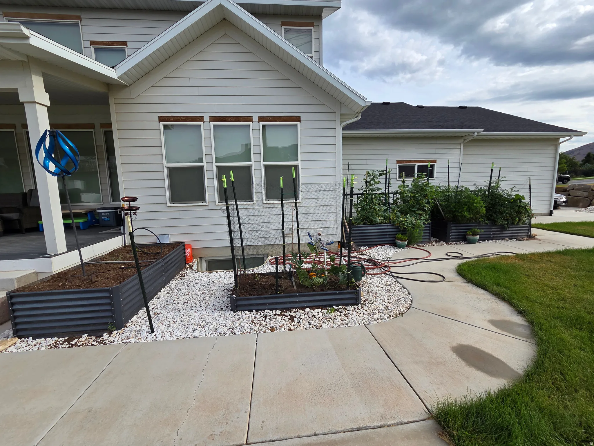Modern home exterior showcasing a concrete patio with clean sight lines, raised garden beds with integrated trellising for vertical planting, white fiber cement siding, and manicured lawn with mature landscaping