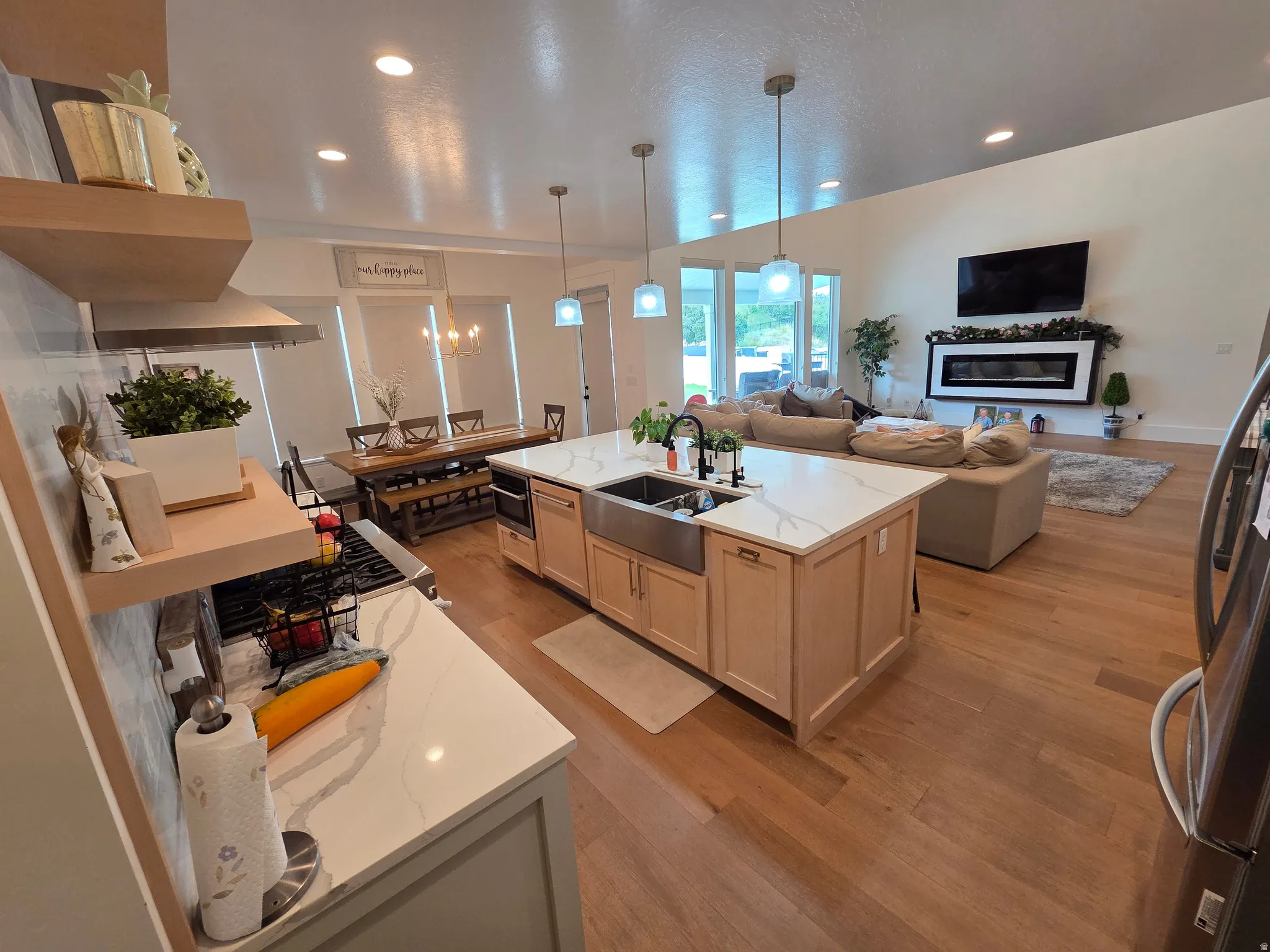 Open-concept kitchen and living area featuring custom wood cabinetry, a large center island with white quartz countertop and farmhouse sink, stainless steel appliances, wood-finish flooring throughout, recessed lighting, and abundant natural light from ex