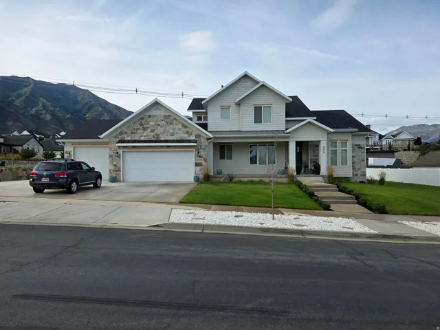 Two-story residence featuring a light-colored exterior with stone and siding accents, a two-car attached garage with a white overhead door, and a manicured front lawn with landscaping