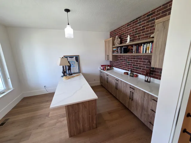Modern home office featuring a wood-finish island countertop with ample workspace, exposed red brick accent wall with built-in shelving and cabinetry, polished concrete countertops, warm wood-finish cabinetry throughout, and pendant lighting