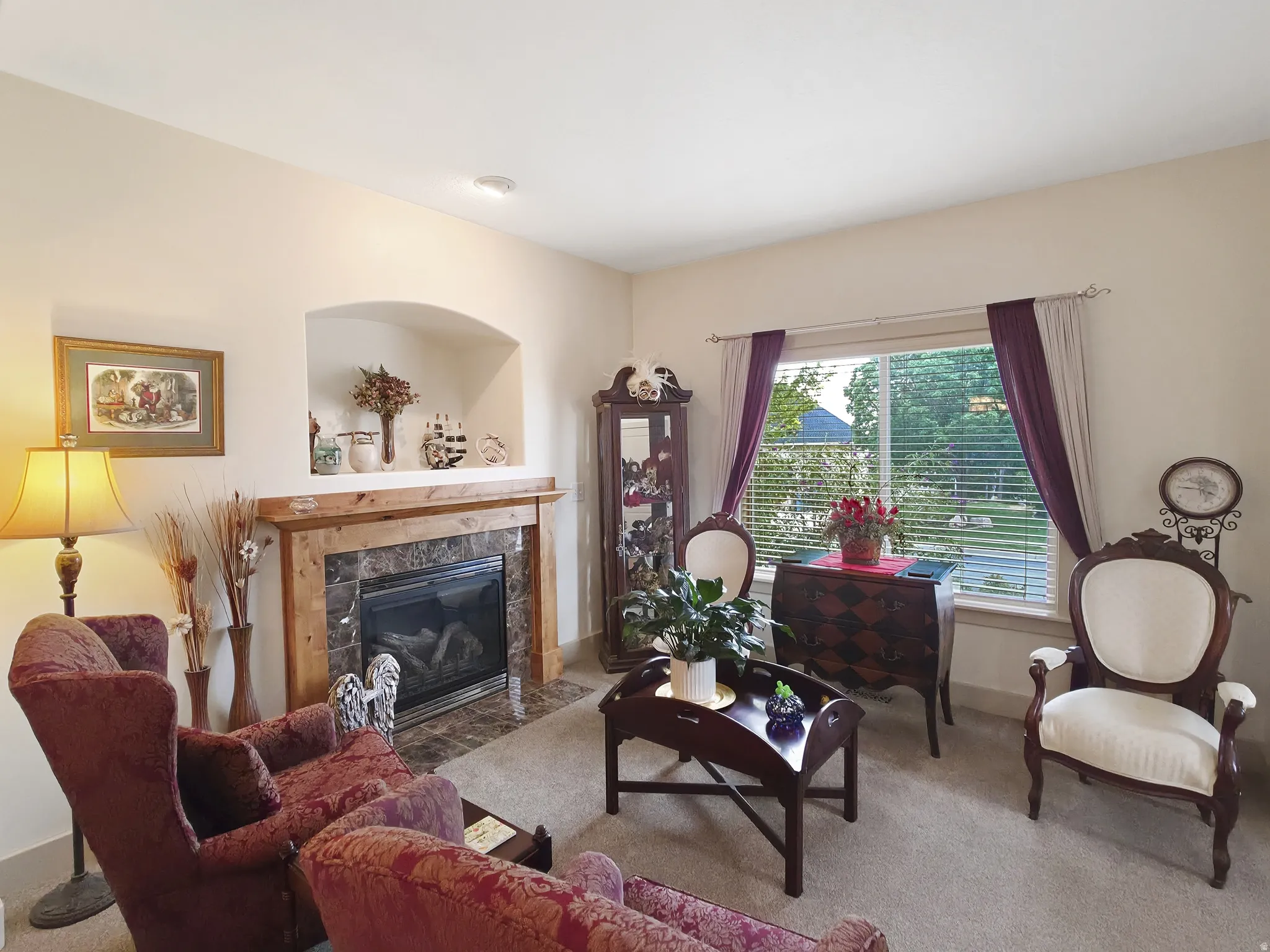 Carpeted living area featuring a fireplace with a wood-finish mantel and stone surround, an arched wall niche, and a window with blinds