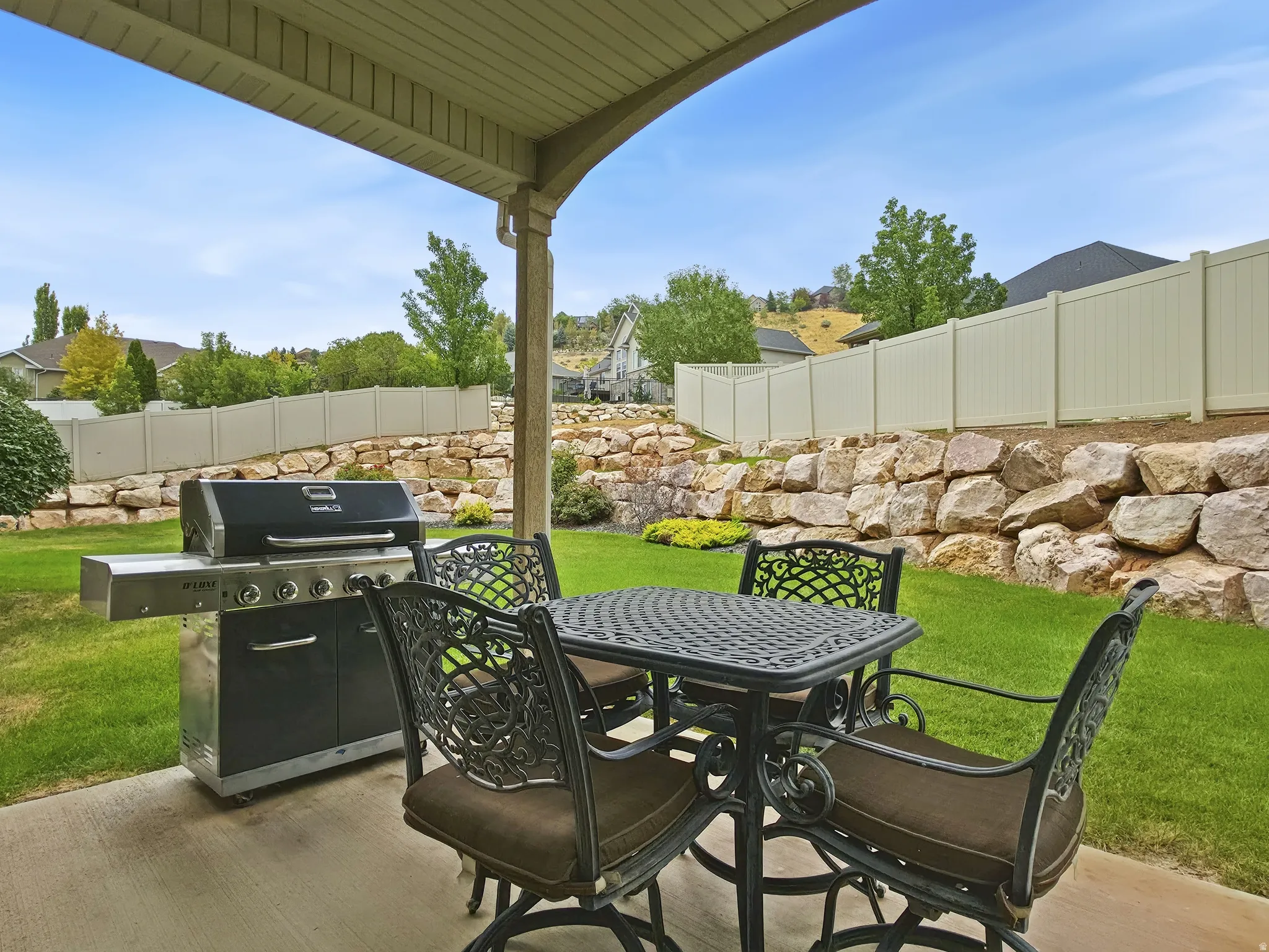 Covered patio with concrete flooring, vibrant green lawn, rock retaining wall, and privacy fencing
