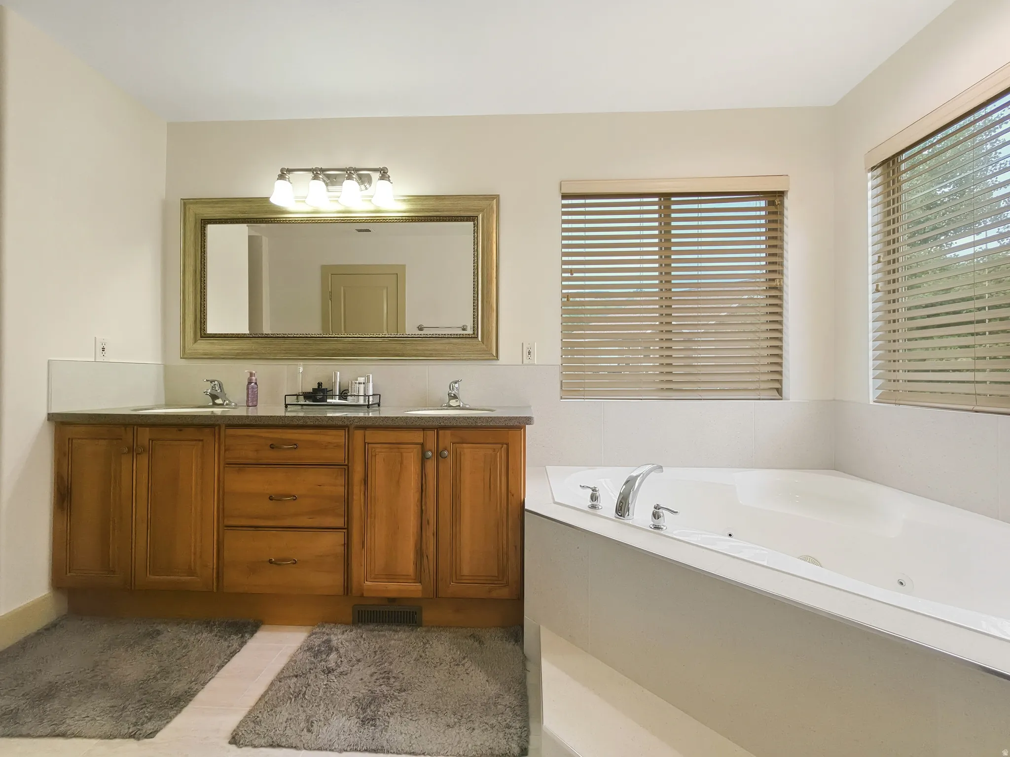 Dual sink vanity with wood-finish cabinetry, corner soaking tub, and multiple windows with horizontal blinds