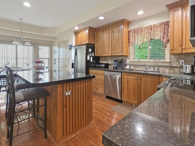 Kitchen featuring wood-finish cabinetry, stainless steel appliances, dark countertops, a tiled backsplash, and recessed lighting