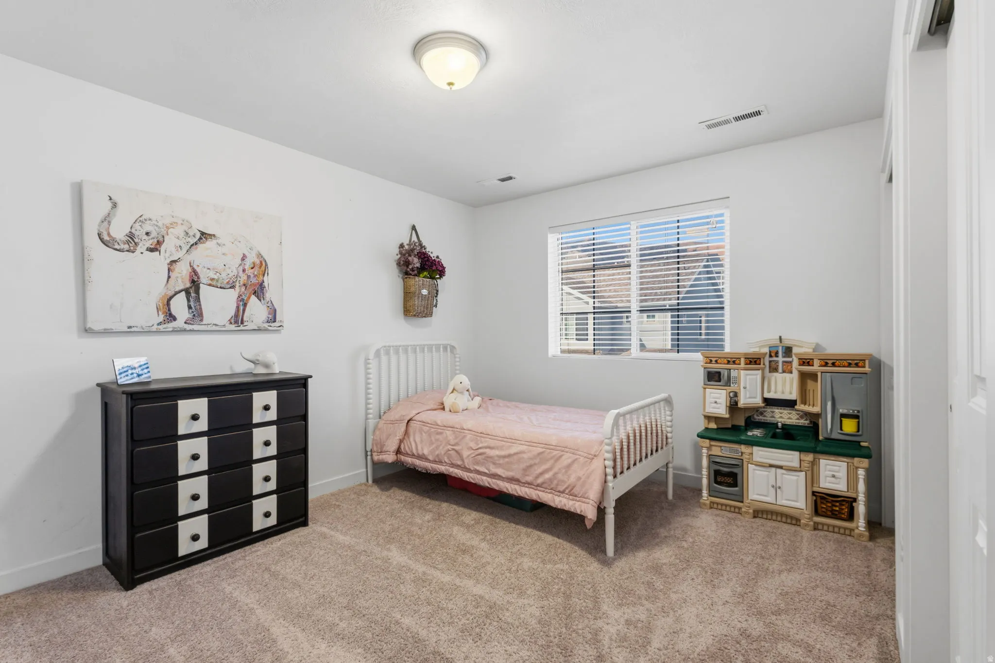 Bright secondary bedroom featuring soft carpet flooring, large windows with plantation shutters, white metal bed frame, and ample storage with wooden cube shelving units