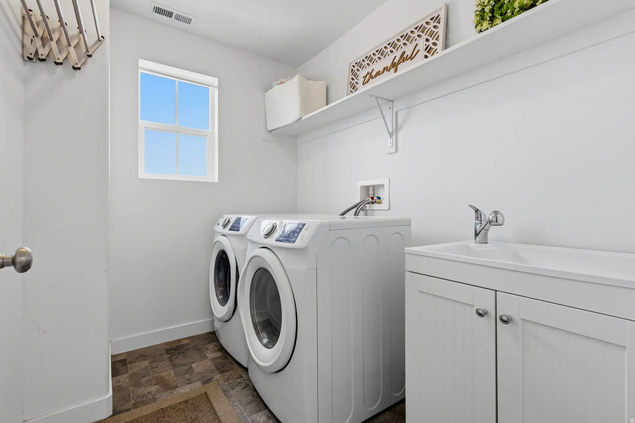 Dedicated laundry room featuring side-by-side front-load washers and dryers, white cabinetry with ample storage, a utility sink with chrome faucet, wood-finish flooring, and a bright window for natural light