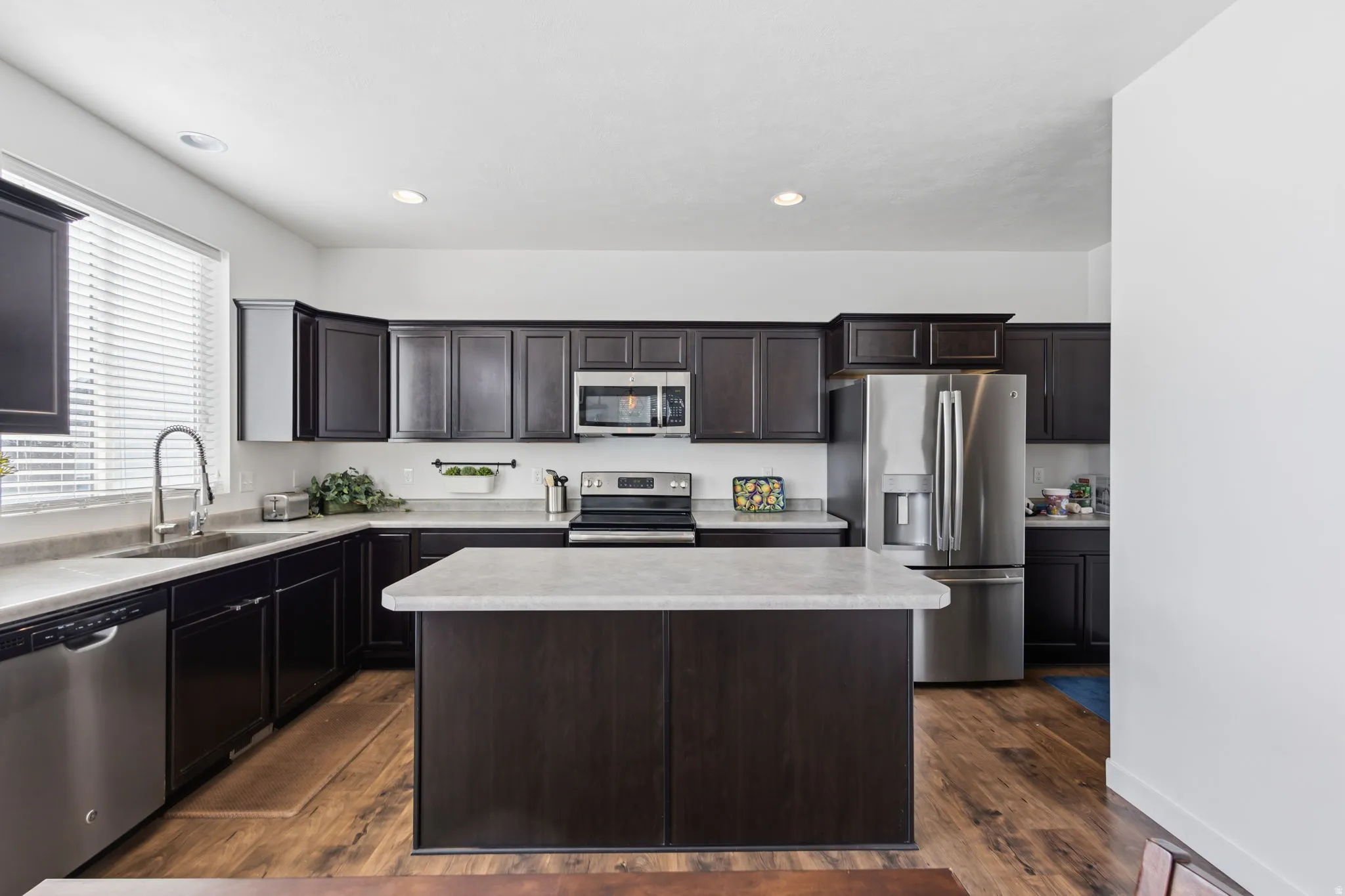 Modern kitchen featuring dark cabinetry with recessed panel detailing, white quartz countertops, a large center island with waterfall edge, stainless steel appliances including a built-in microwave and refrigerator, and warm wood-finish flooring throughou