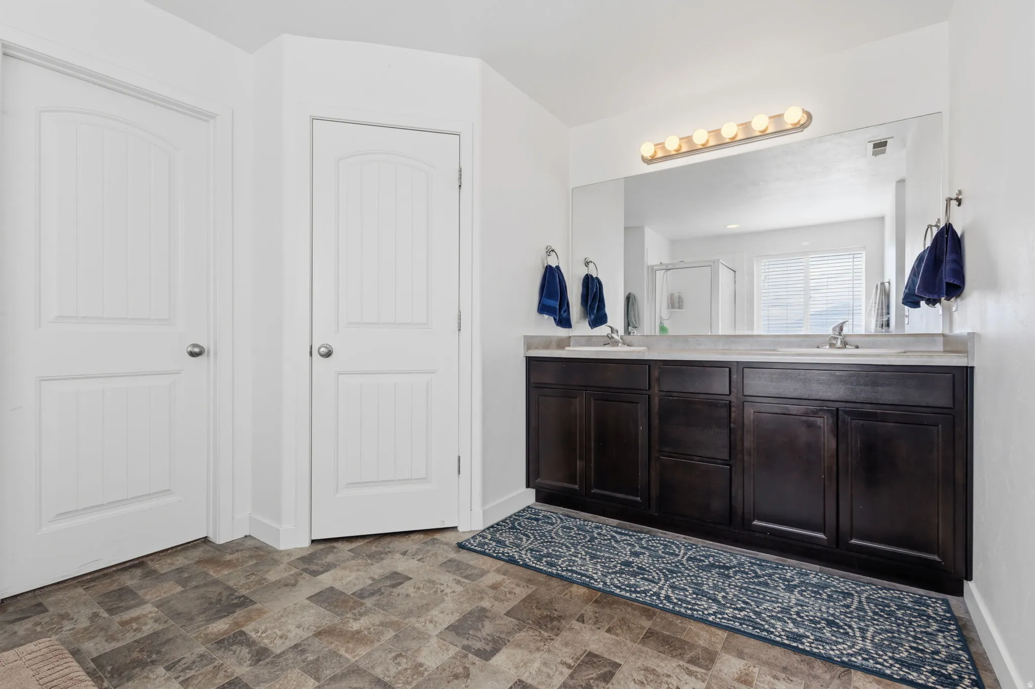 Spa-inspired primary bathroom featuring a dark wood double vanity with dual sinks, extensive mirror with integrated vanity lighting, tile flooring, and dual white six-panel closet doors for ample storage