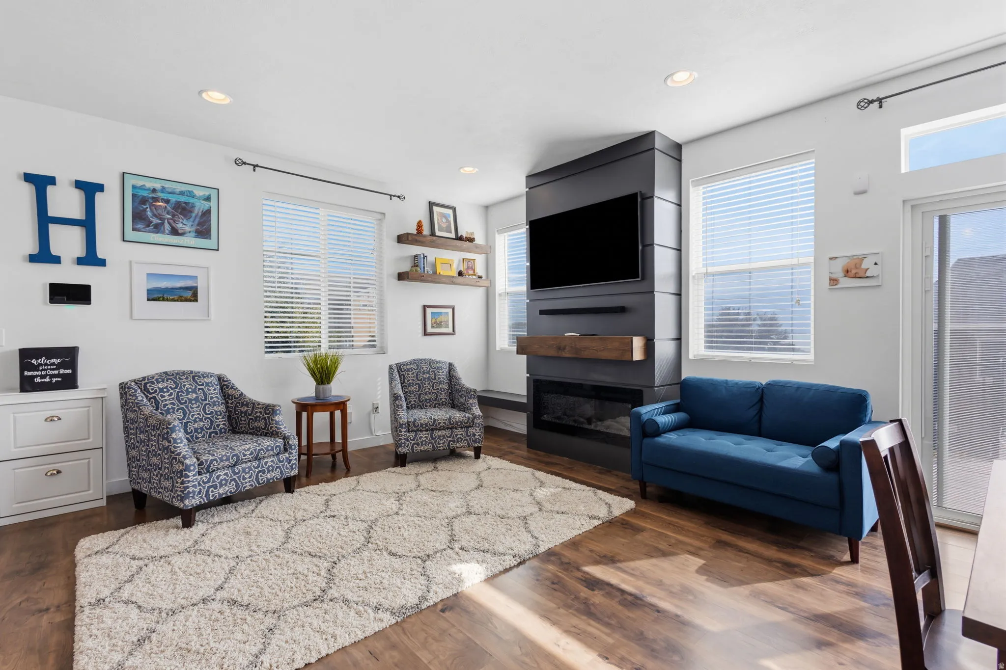 Bright and sophisticated living space featuring hardwood flooring, a gas fireplace with stone surround and built-in shelving, a blue upholstered sofa, patterned accent chairs, and expansive windows with plantation shutters that flood the room with natural