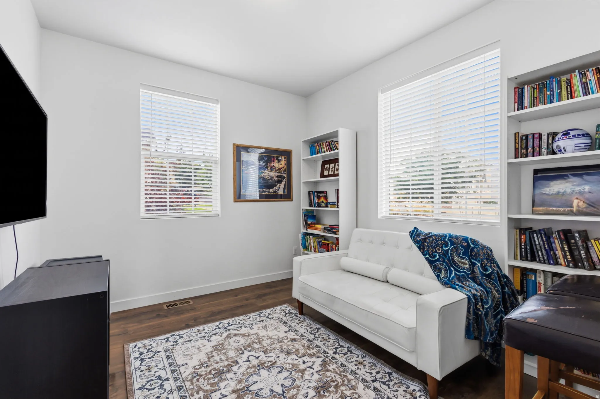 Bright, light-filled media room featuring multiple windows with plantation shutters, hardwood flooring, built-in shelving throughout, and a flat-screen television mounted on the wall