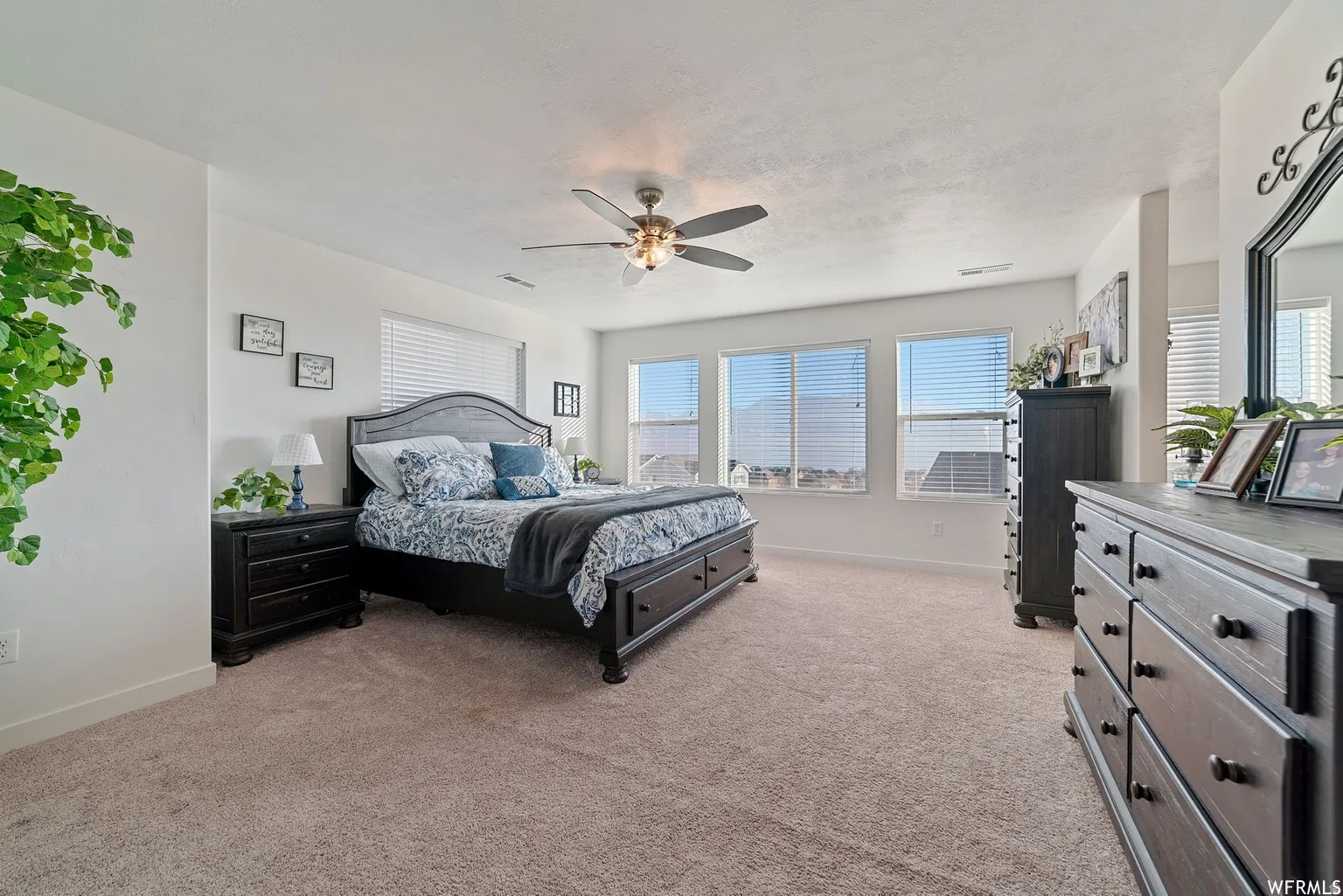 Spacious primary bedroom suite showcasing multiple large windows with expansive natural light, neutral carpet flooring, ceiling fan with integrated lighting, black wood-finish bedroom furniture, and a coordinating dresser with mirror