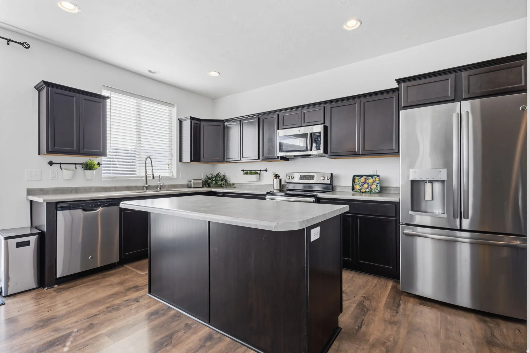 Modern kitchen featuring dark cabinetry with soft-close doors, a large waterfall-edge island with seating, quartz countertops, stainless steel appliances including a French-door refrigerator and built-in microwave, and wood-finish flooring throughout the