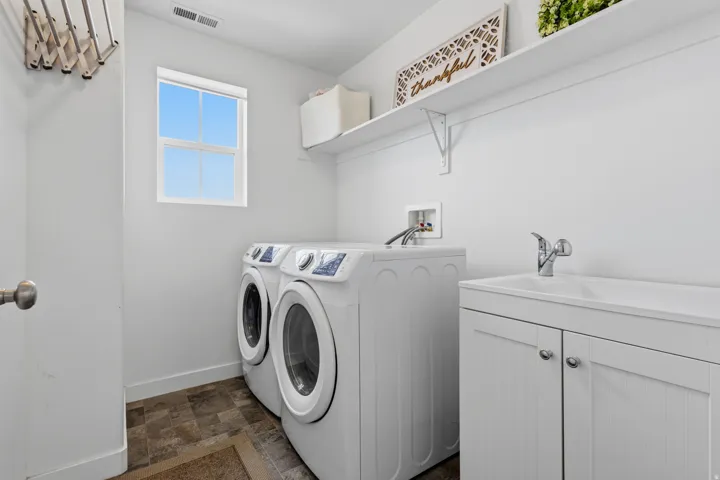 Dedicated laundry room featuring side-by-side front-load washers and dryers, white cabinetry with ample storage, a utility sink with chrome faucet, wood-finish flooring, and a bright window for natural light