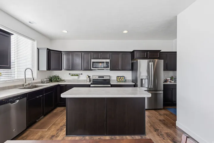 Modern kitchen featuring dark cabinetry with recessed panel detailing, white quartz countertops, a large center island with waterfall edge, stainless steel appliances including a built-in microwave and refrigerator, and warm wood-finish flooring throughou