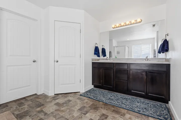 Spa-inspired primary bathroom featuring a dark wood double vanity with dual sinks, extensive mirror with integrated vanity lighting, tile flooring, and dual white six-panel closet doors for ample storage