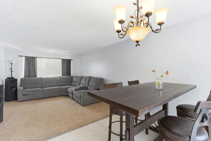 Open living and dining space anchored by a dark wood-finish dining table, neutral-toned sectional sofa, decorative chandelier with multiple glass shades, large windows with gray drapery, and warm wood-finish flooring throughout