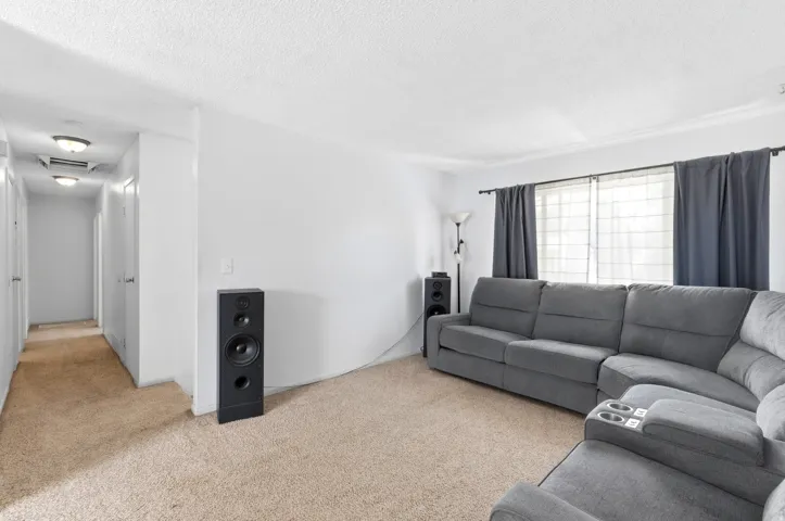 Spacious living area featuring neutral carpeting, large windows with dark curtains, and a wall-mounted speaker system