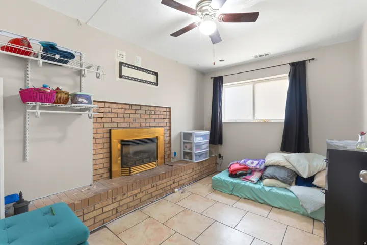 Spacious recreation room featuring a brick fireplace with yellow surround, tile flooring, ceiling fan with integrated lighting, wall-mounted shelving, and large windows with blackout drapery