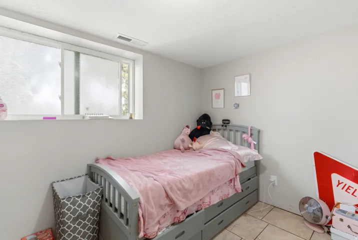 Bright secondary bedroom featuring tile flooring, large window with natural light, gray painted wood bed frame with integrated storage drawers, and built-in wall shelving for storage and display