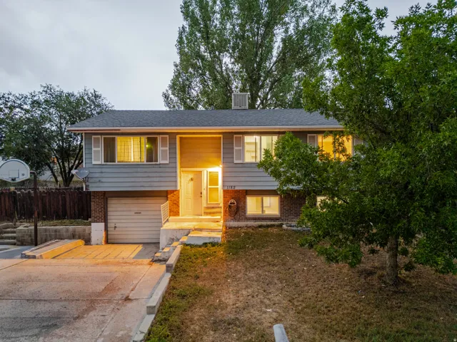 Two-story residence featuring a brick and siding exterior with a two-car garage, illuminated entry vestibule with warm lighting, and mature landscaping including established trees flanking the property