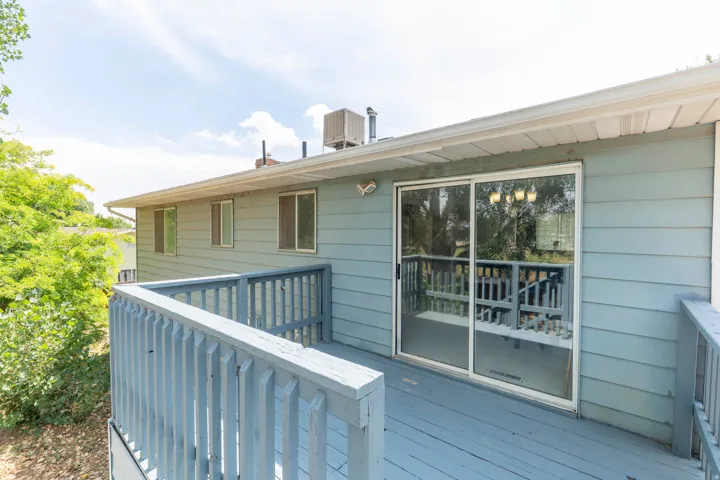 Covered deck with white aluminum railings and horizontal slatted balusters, large sliding glass doors with reflective panes, horizontal fiber cement siding in soft blue-gray, and mature landscaping with established greenery framing the exterior
